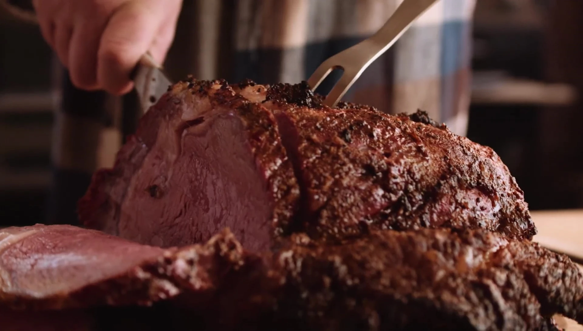 The meat fork holding a roast in place on a cutting board.